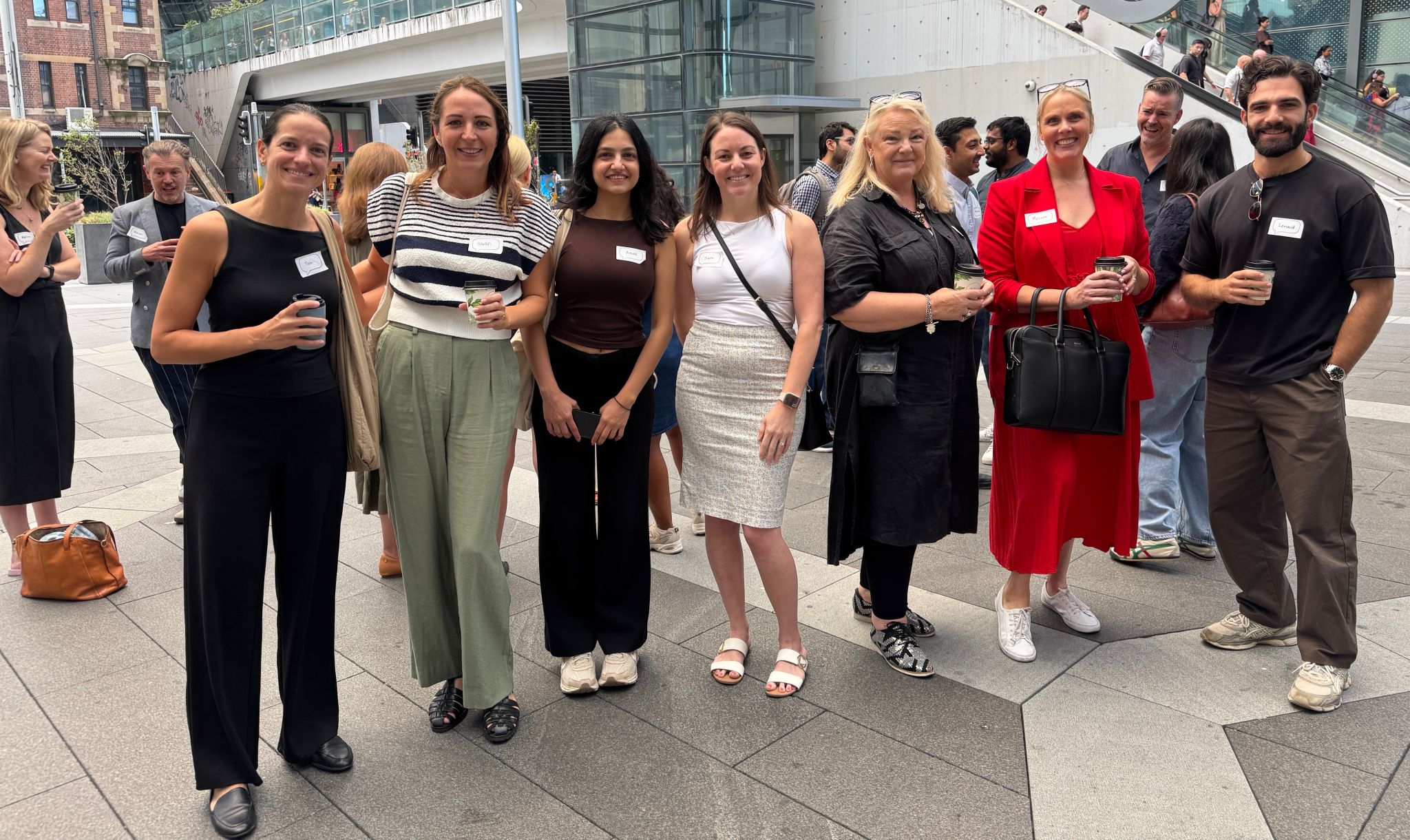 A group of people standing in a rough line, looking to camera. Some are carrying a coffee, all have name tags