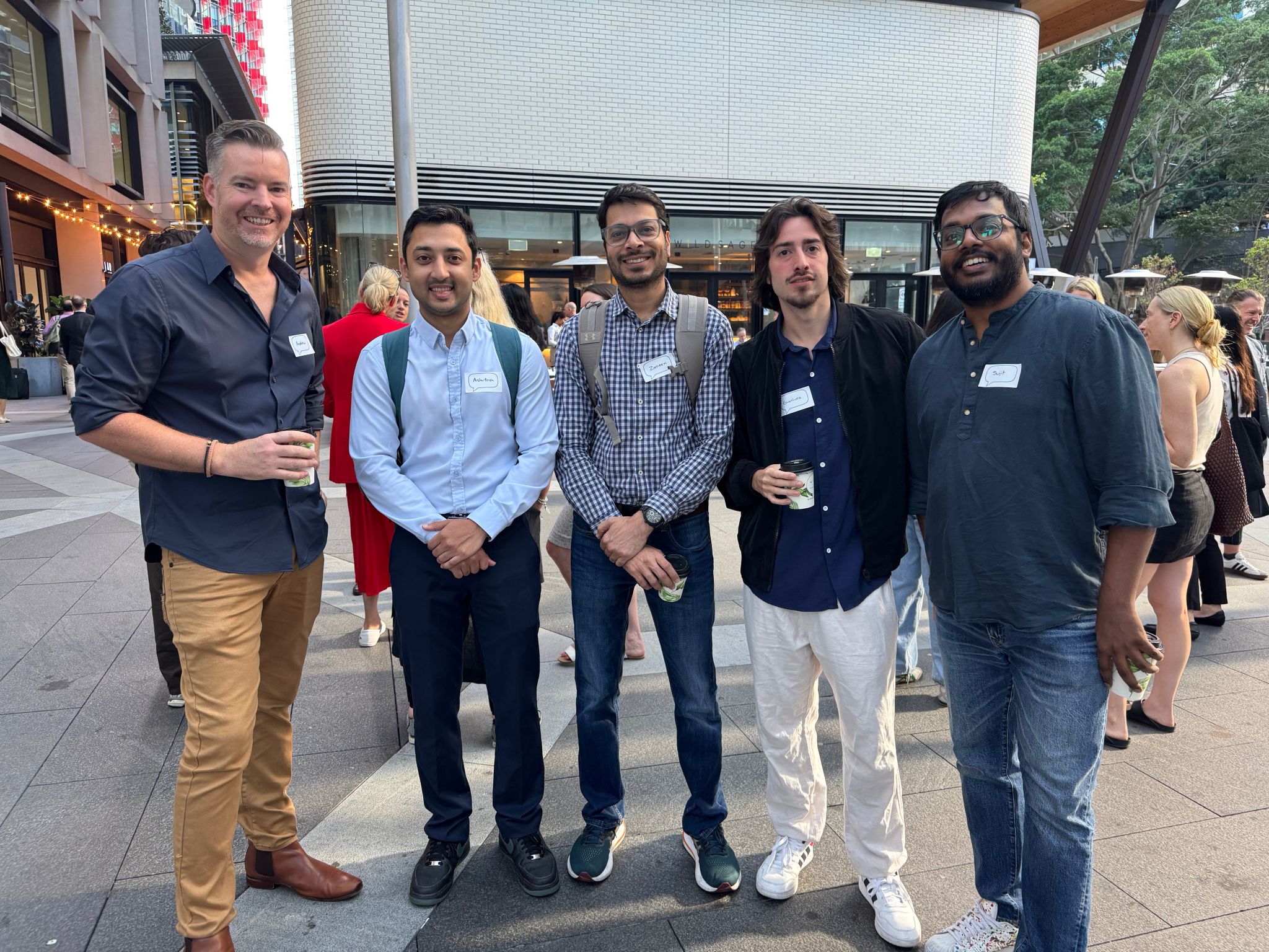 A group of people standing in a rough line, looking to camera. Some are carrying a coffee, all have name tags