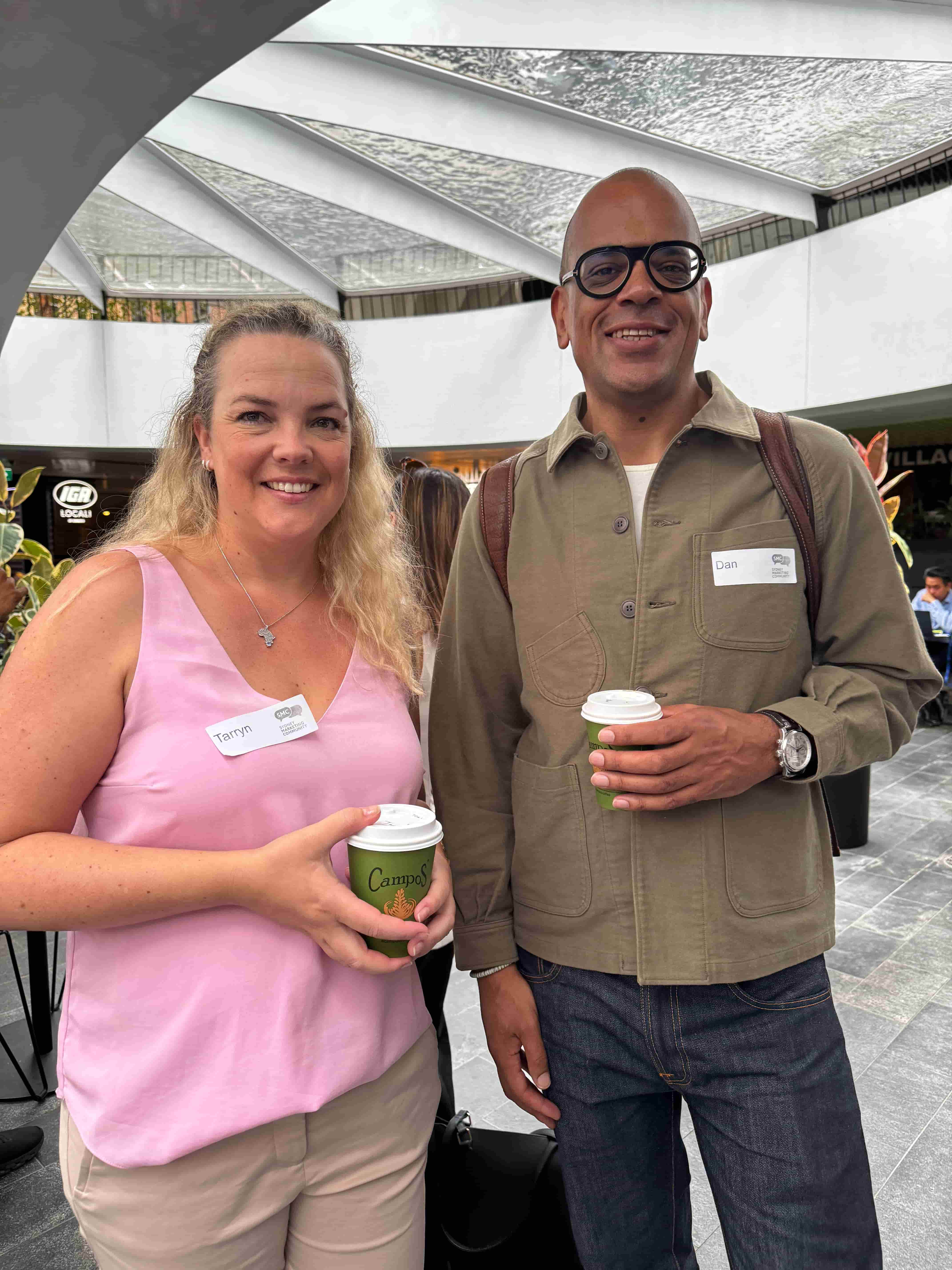 A group of people standing in a rough line, looking to camera. Some are carrying a coffee, all have name tags