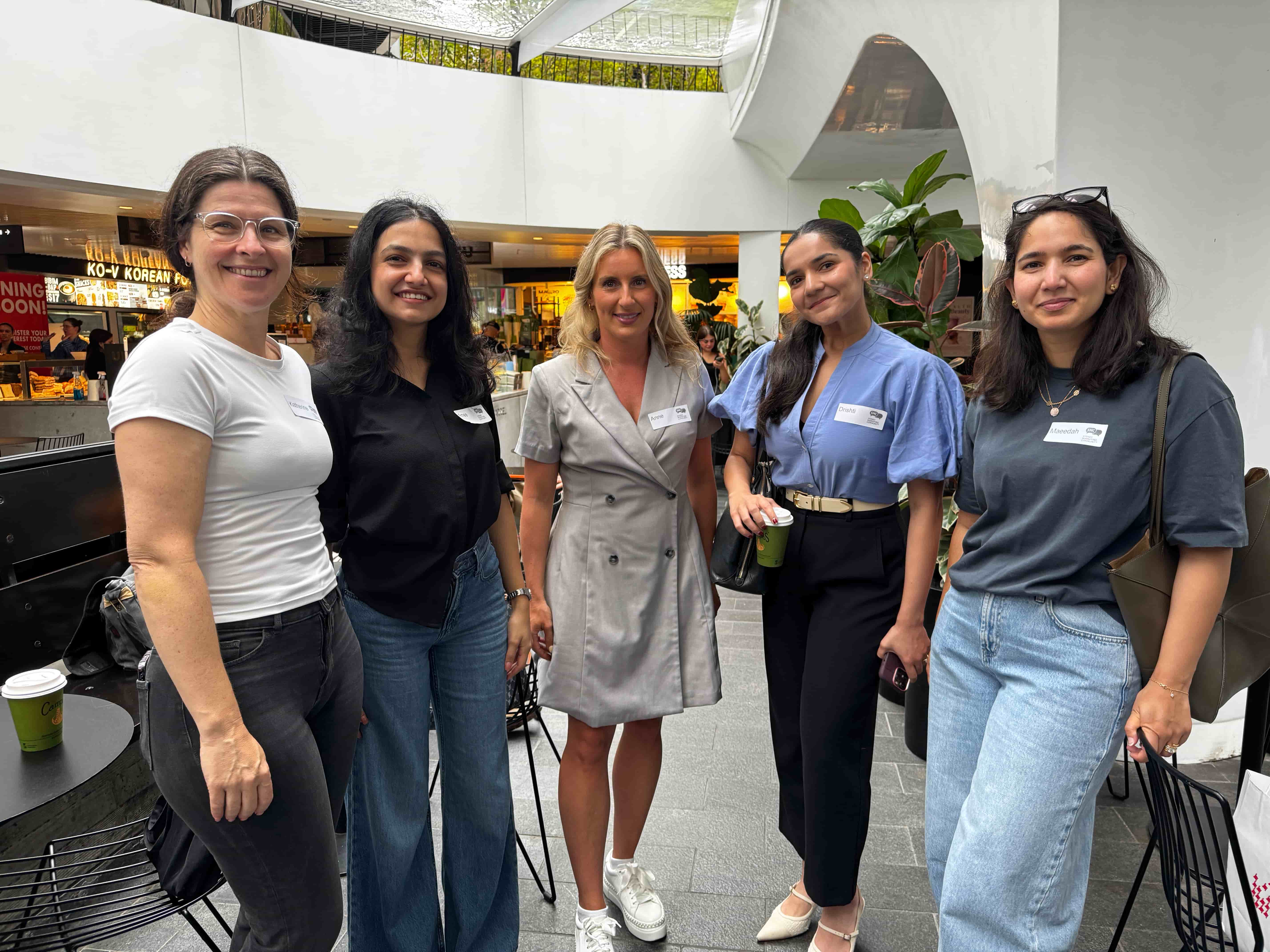 A group of people standing in a rough line, looking to camera. Some are carrying a coffee, all have name tags
